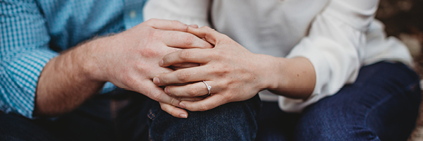 Couple sit together holding hands, woman wears engagement ring.